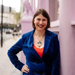 Kathryn, a white woman with chin length brown hair, in front of a nearly black wall background. Kathryn is wearing a royal blue suit, bright orange top & frida kahlo necklace.