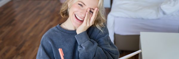 Portrait of happy, young blond woman, sitting in bedroom with notebook and pen, laughing and smiling, writing in journal, doing homework and smiling.